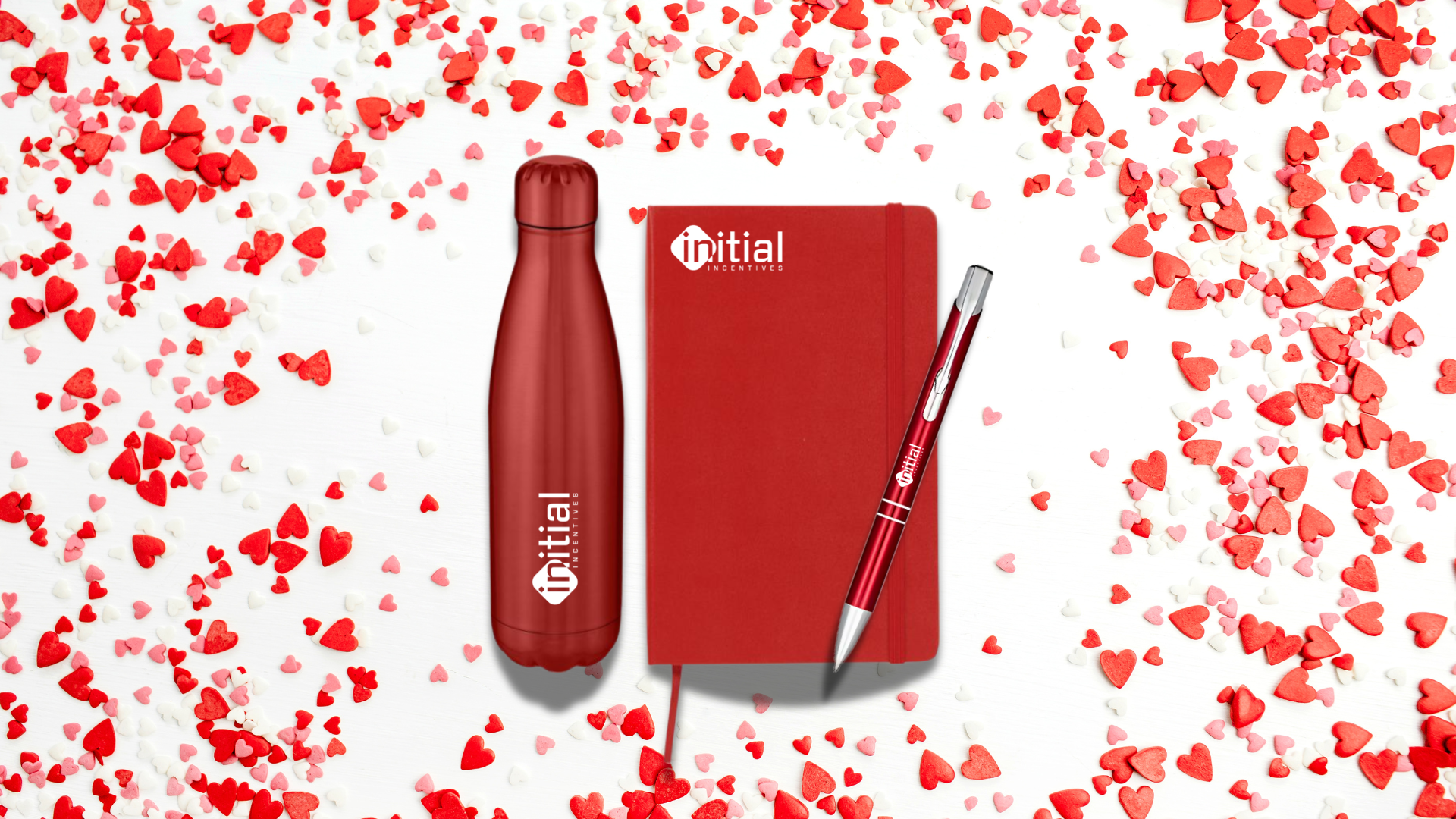 A branded water bottle, printed notebook and engraved pen laying on a table covered in heart confetti.