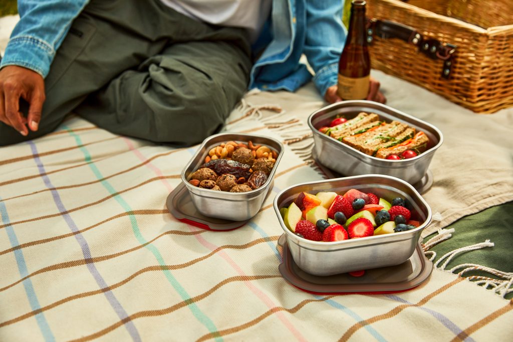 Branded Black+Blum meal prep boxes with 3 different sizes each with their own customisable area, sat on a picnic blanket in the summer evening sun.