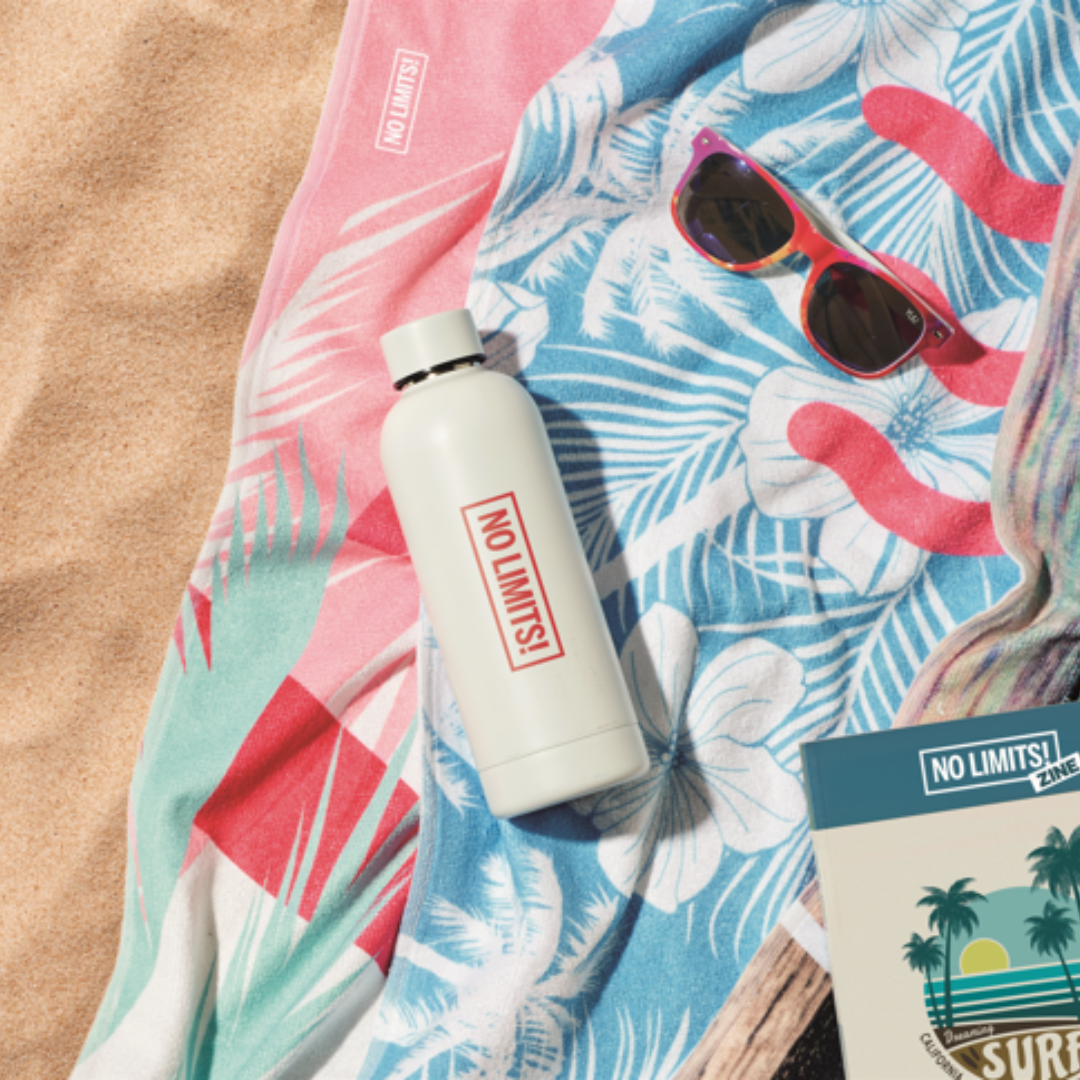 Branded summer merchandise on sand including a NO LIMITS! water bottle, beach towel, sunglasses, and zine.