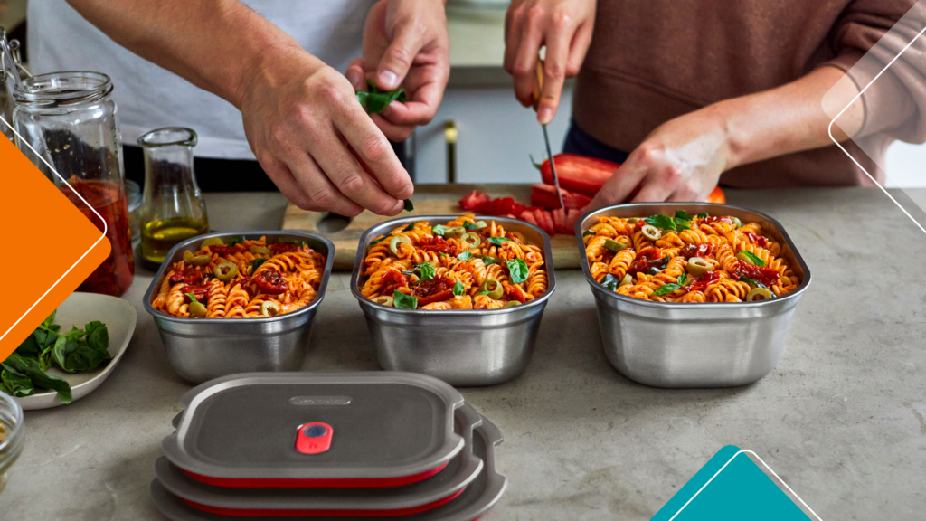 People preparing three stainless steel meal prep containers filled with pasta, olives and fresh herbs on a kitchen worktop.